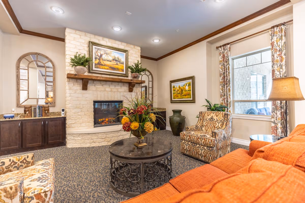 A cozy living room with a stone fireplace, a round coffee table with a floral arrangement, an orange sofa, and patterned armchairs. The room features two framed paintings, decorative plants, a lamp, and a window with floral curtains.