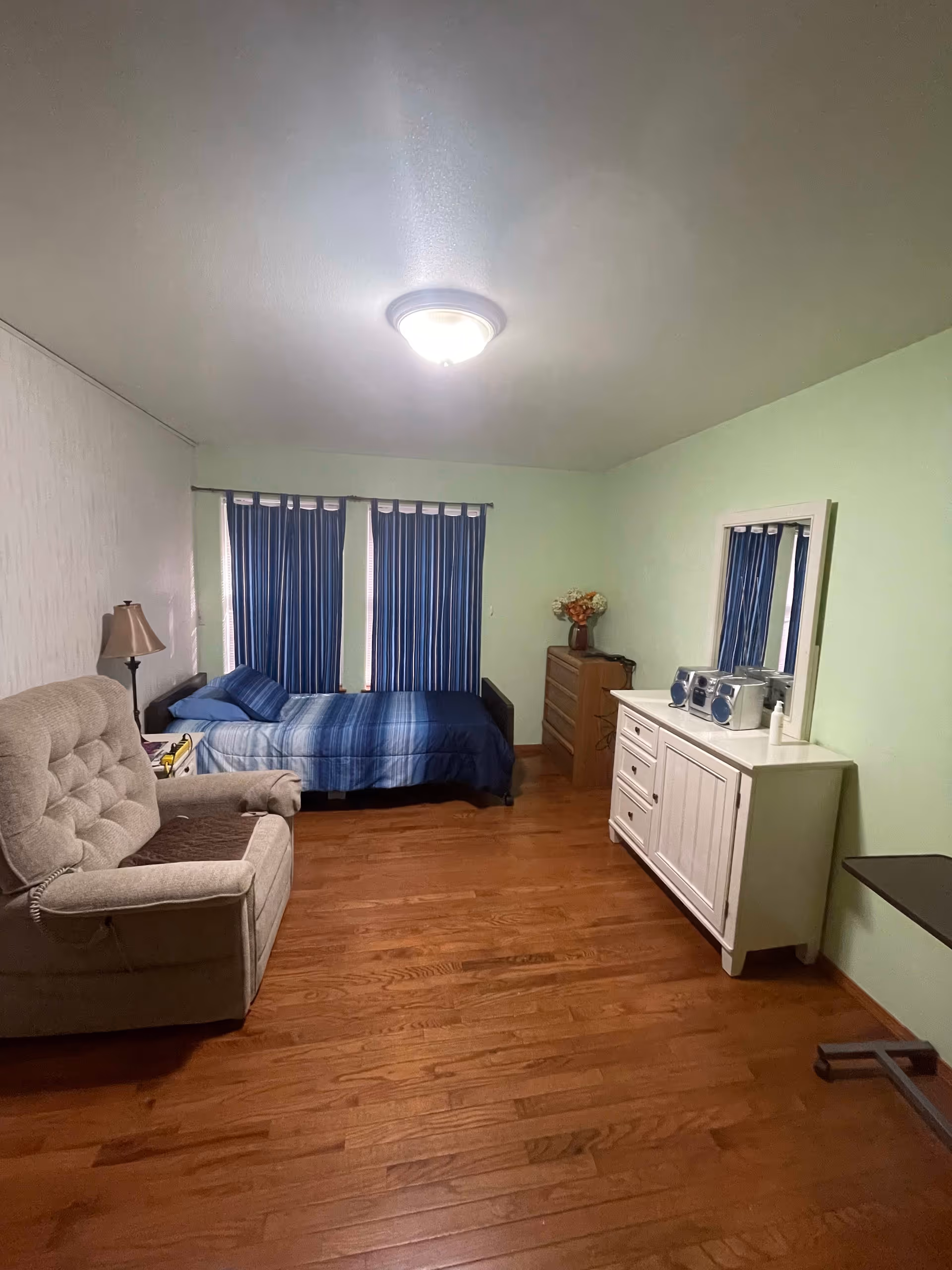 A bedroom with wooden flooring, a single bed with blue striped bedding against the wall with two windows covered by blue curtains. There is a beige armchair with a brown cushion on the left side, a floor lamp next to it, a white dresser with a mirror and a stereo on top on the right side, and a wooden chest of drawers with a vase of flowers in the corner.