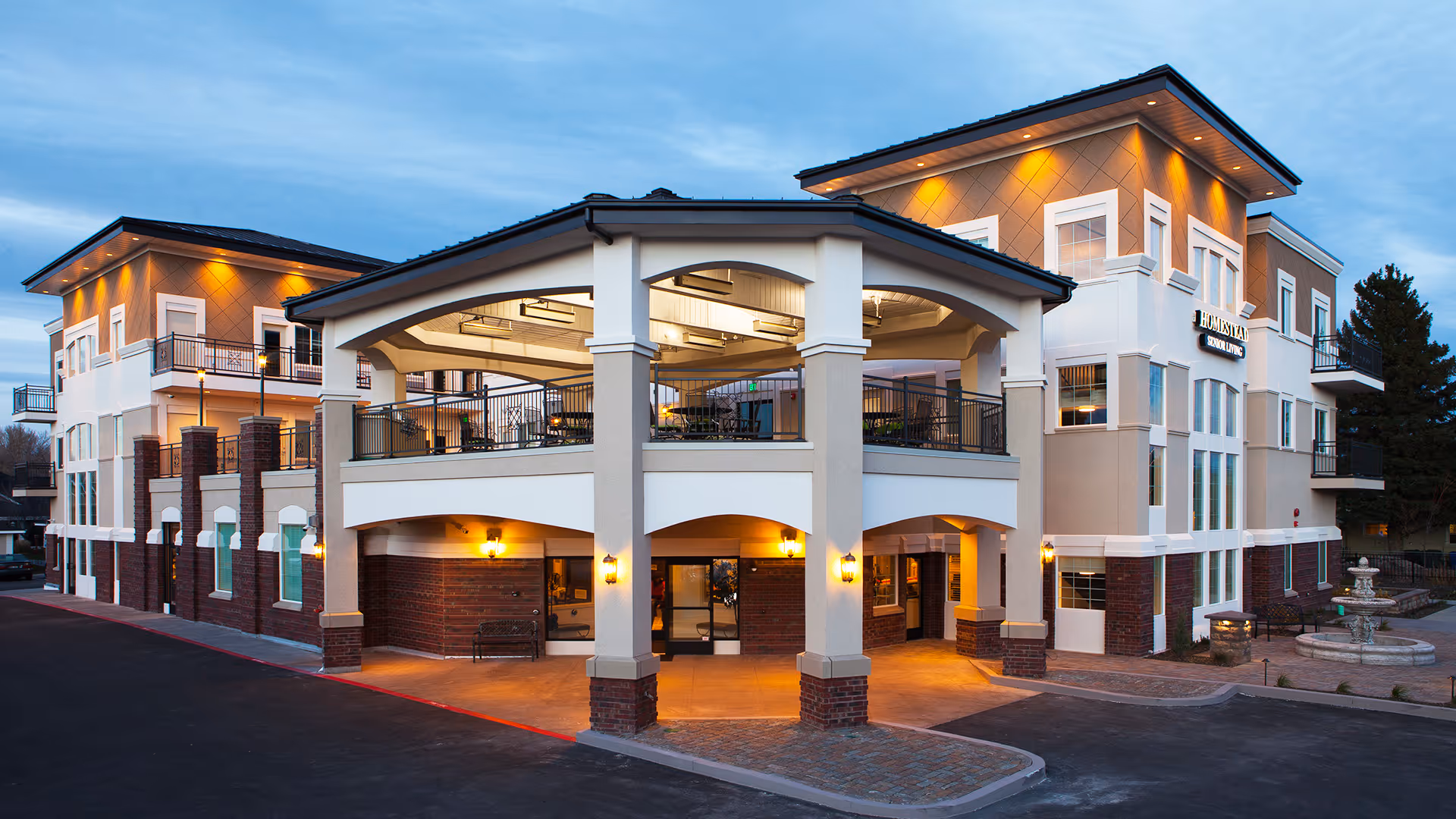 Exterior view of Homestead Assisted Living facility during dusk, showing a modern three-story building with illuminated outdoor seating area on the second floor, warm lighting, and a fountain on the right side.