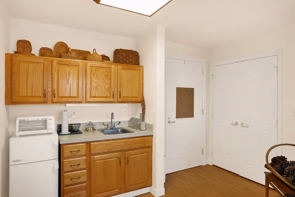 Small kitchenette area with wooden cabinets, a countertop with a sink, a mini refrigerator, a toaster oven, and various baskets on top of the cabinets. There are two white doors on the right side of the kitchenette, and a basket with pinecones is partially visible in the bottom right corner.
