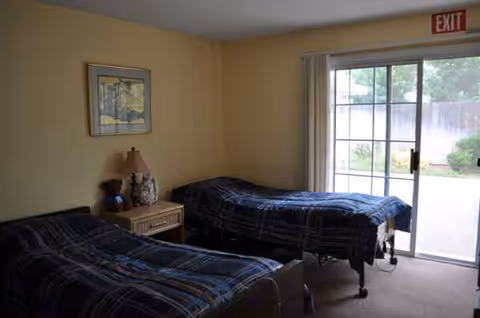 A modest bedroom features two twin beds, a nightstand, lamp, painting, and sliding glass door.