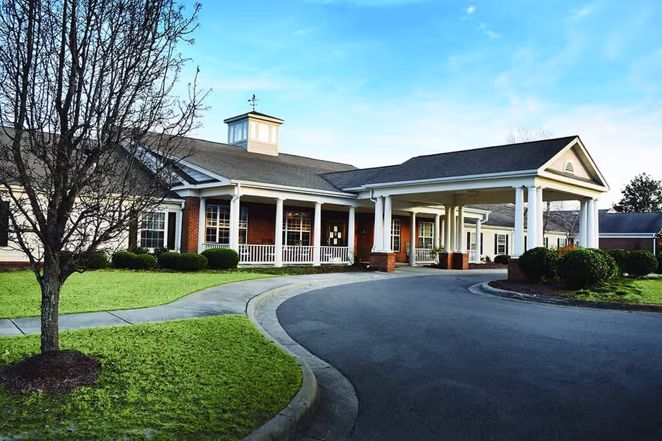 Exterior view of a single-story senior living facility building with a covered entrance supported by white columns. The building has a brick facade with white trim and multiple windows. There is a curved driveway leading to the entrance and a small tree and bushes in the landscaped grassy area in front.