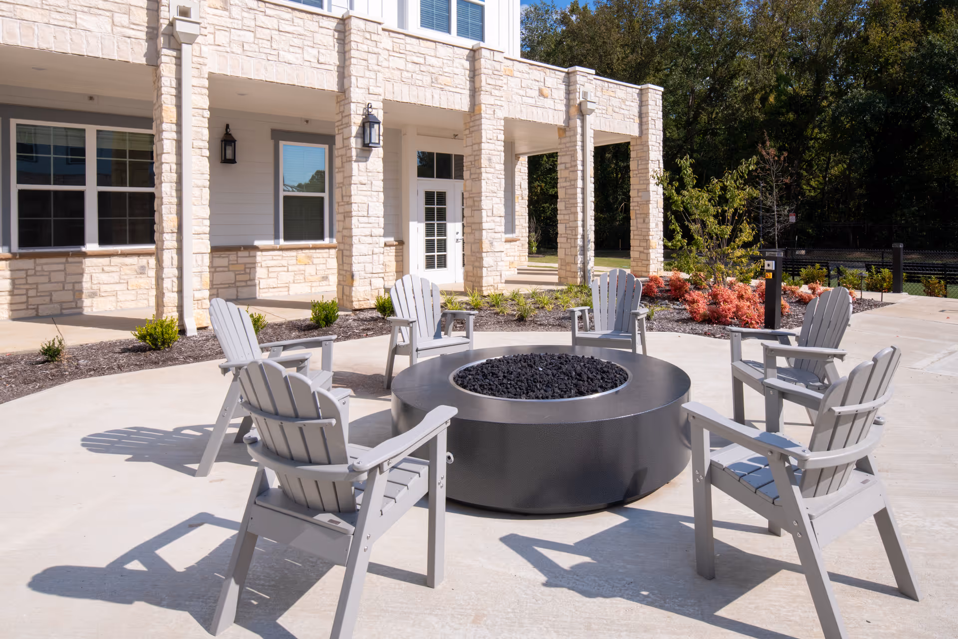 Outdoor patio area at The Summit of Germantown featuring six gray Adirondack chairs arranged in a circle around a large round fire pit. The background shows a stone and siding building facade with windows, doors, and outdoor wall lanterns. There are landscaped plants and trees surrounding the patio area under a clear sky.