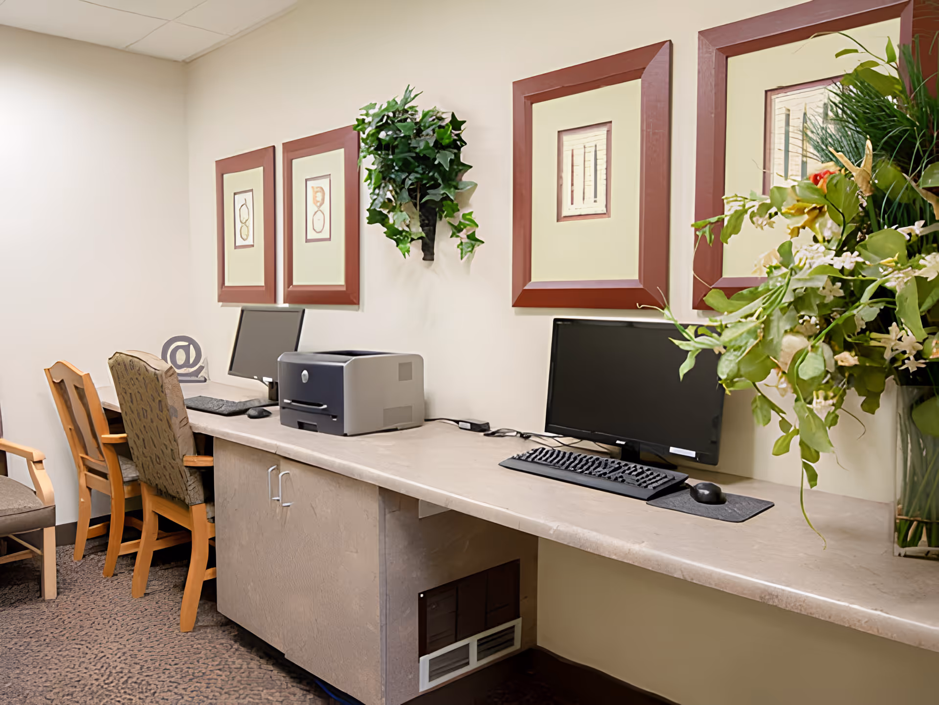 A small office or computer area with two desktop computers, a printer, and three chairs. The wall above the desk has four framed pictures and a decorative green plant. There is also a large floral arrangement on the right side of the desk.