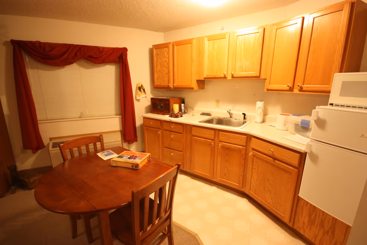 Small kitchen with wooden cabinets, a sink and refrigerator, and a dining table with two chairs beneath a window with red curtains.