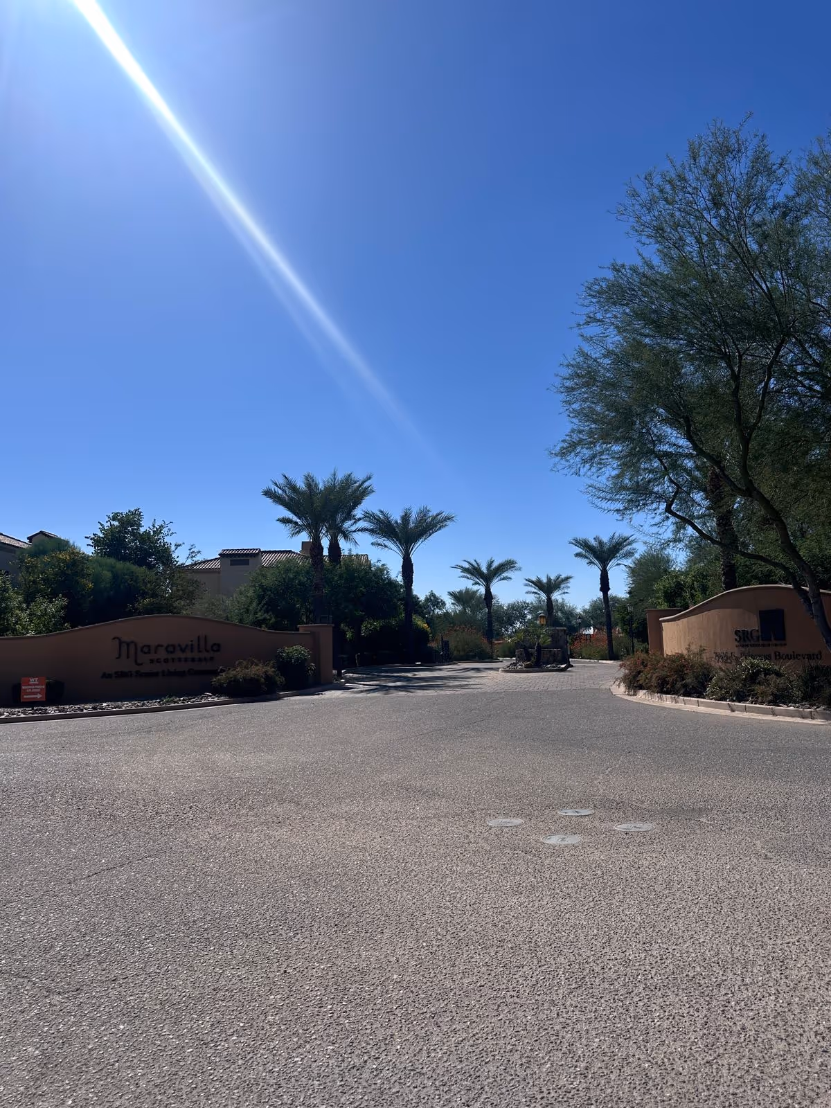 Driveway entrance to Maravilla Scottsdale with palm trees, landscaped walls and a clear blue sky.