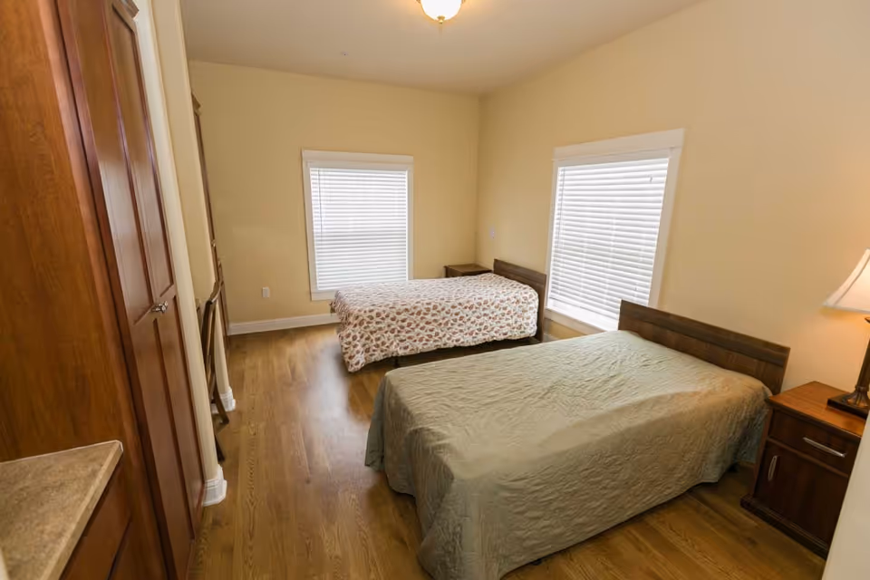 A simple bedroom with two single beds, wooden furniture, hardwood floors and two windows with blinds.