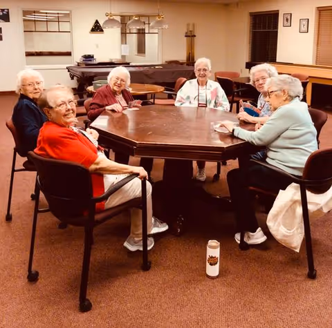 Six elderly women sitting around a large wooden octagonal table in a well-lit room, engaging in a card game. The room has carpeted floors, several chairs, a pool table in the background, and framed pictures on the walls.