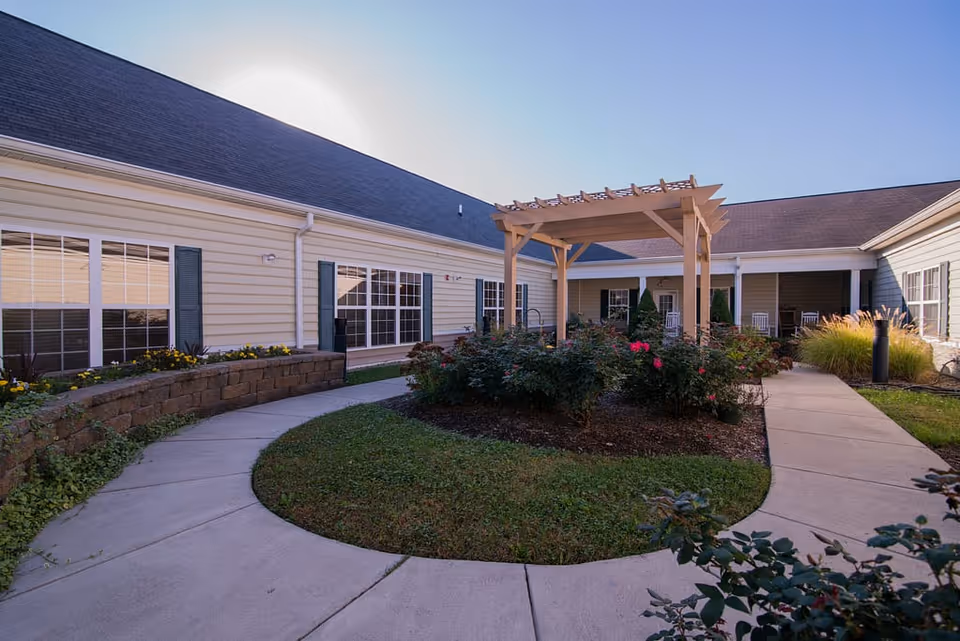 Sunlit courtyard with a wooden pergola, circular walkways, flower beds, and a surrounding single-story building.
