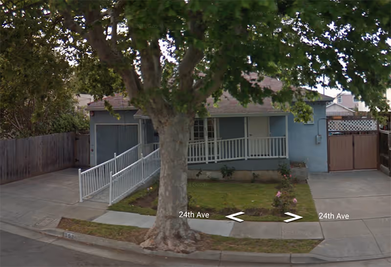 Front exterior view of a single-story blue building with a ramp leading to the entrance, a large tree in the front yard, and a driveway on either side. The sidewalk and street are visible with '24th Ave' street markings.