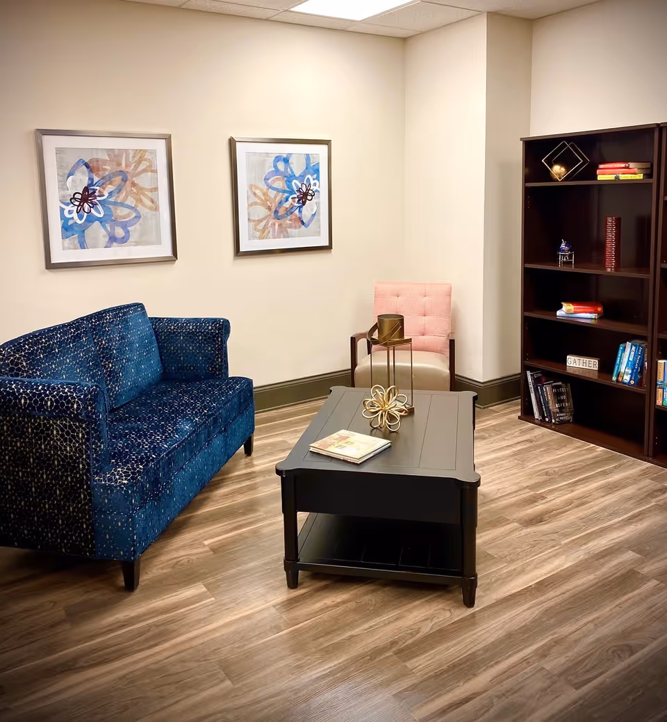 A cozy living room area with a blue patterned loveseat, a pink cushioned chair, a black wooden coffee table with decorative items and a book on it, and a dark wooden bookshelf filled with books and decorative pieces. Two framed abstract floral artworks hang on the beige wall above the loveseat.