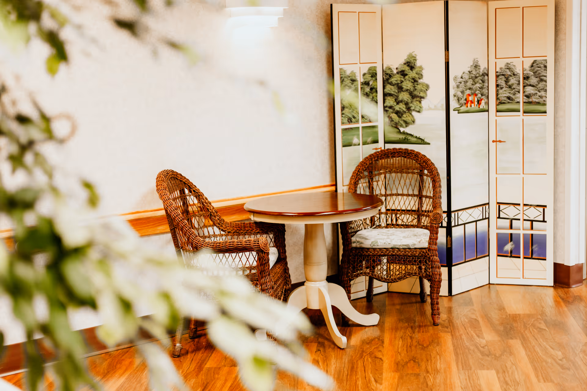 A small seating area with two wicker chairs with cushions and a round wooden table between them, placed on a wooden floor. Behind the chairs is a decorative folding screen with painted landscape scenes. Some green foliage is partially visible in the foreground.