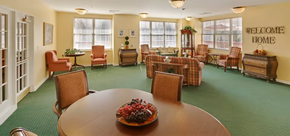 A cozy living room area with green carpet and yellow walls. The room features multiple armchairs and sofas with plaid upholstery, a round wooden table with chairs, and a decorative bowl of grapes on the table. Large windows allow natural light to fill the space. On the right wall, there is a decorative sign that reads 'WELCOME HOME'. Various pieces of wooden furniture and plants add to the warm ambiance.