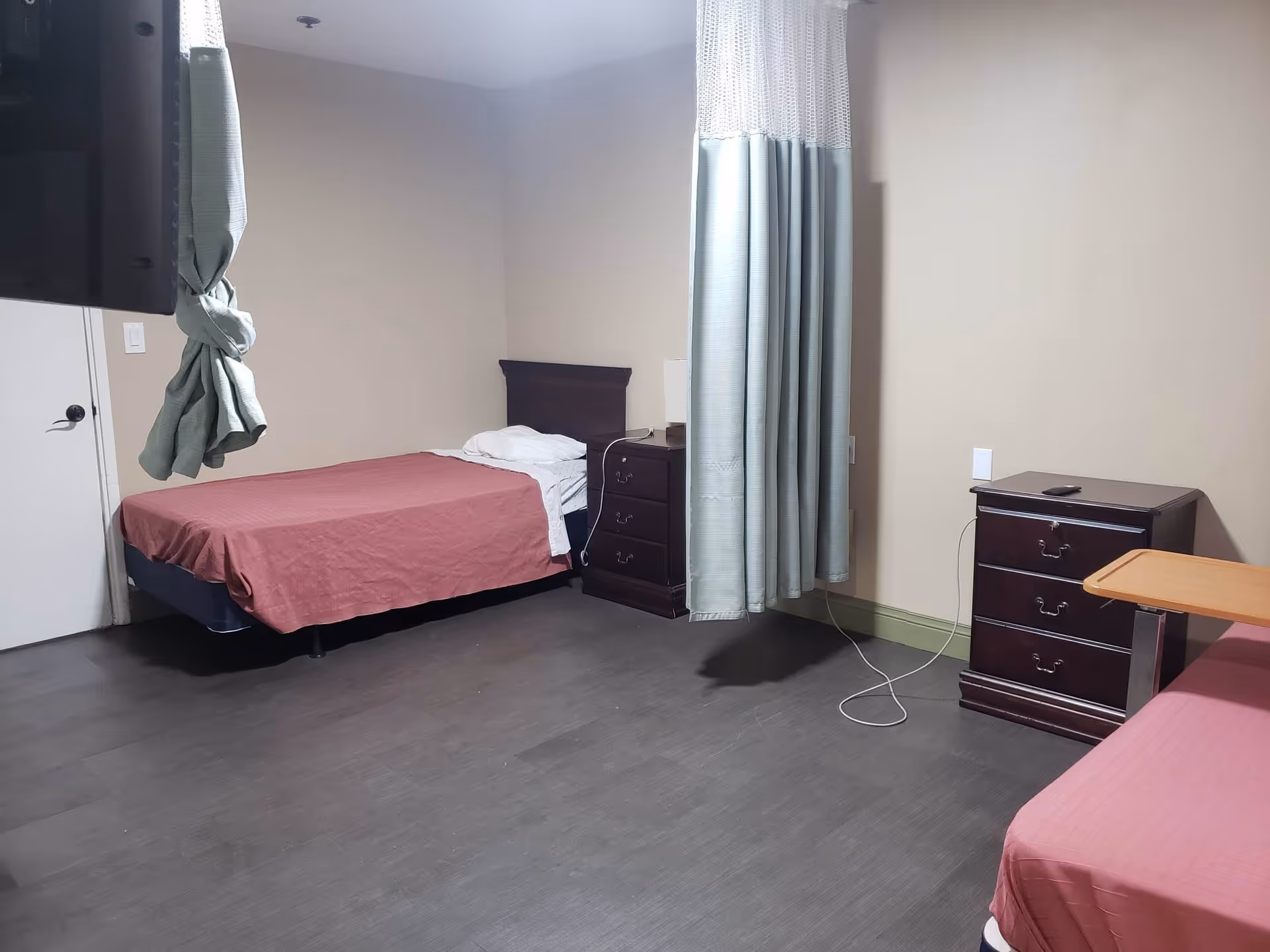 A simple bedroom in a senior living facility with two single beds covered in red bedspreads, a dark wooden nightstand with a lamp between the beds, a curtain partially drawn in the middle of the room, a small dark wooden dresser with a remote on top, and a wall-mounted TV on the left side.