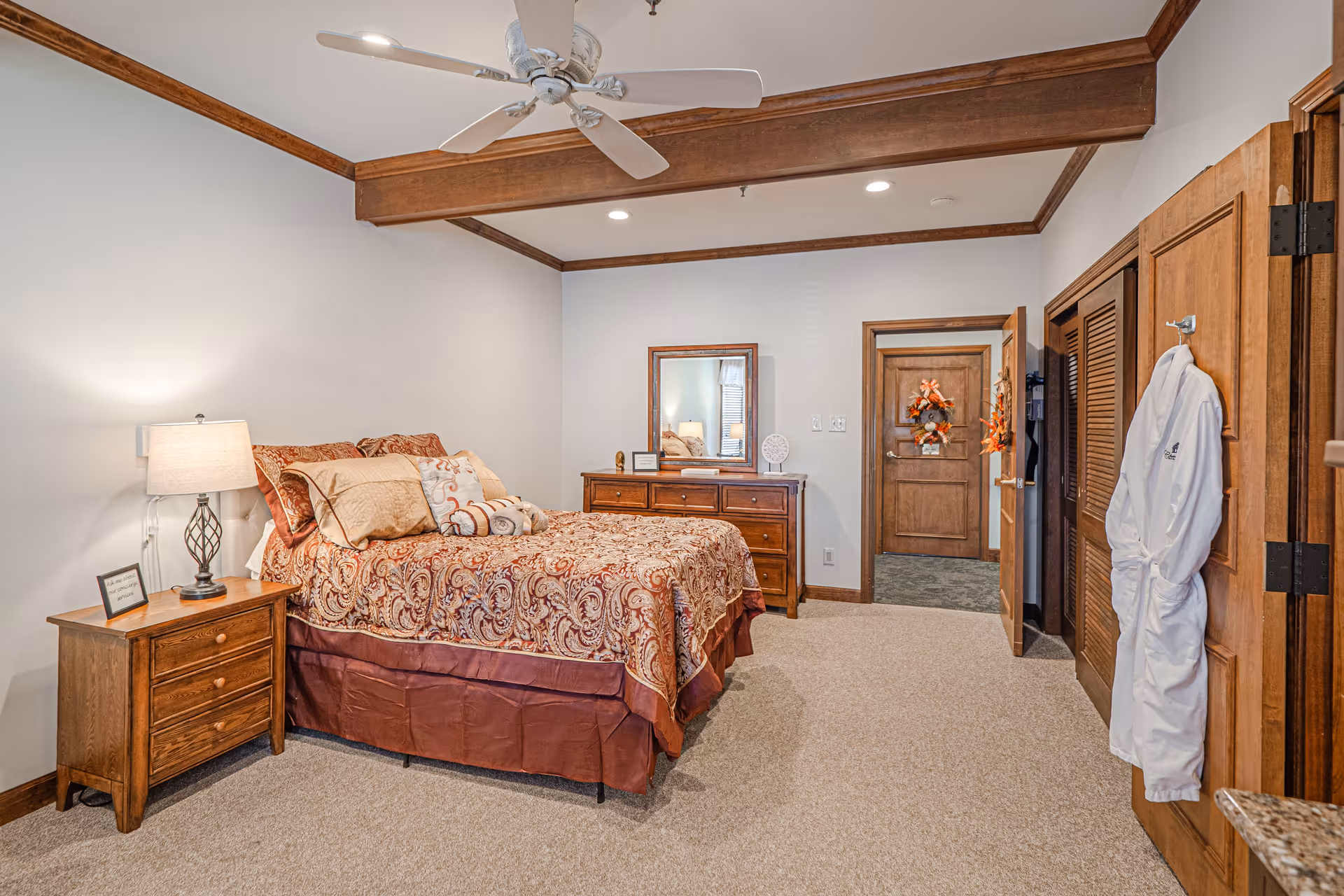 A cozy bedroom with a bed covered in a patterned bedspread and multiple pillows. There is a wooden nightstand with a lamp and a small framed picture on it. Across from the bed is a wooden dresser with a mirror above it. The room has beige carpet, white walls with wooden trim, a ceiling fan, and a white robe hanging on a hook on the wooden closet door.