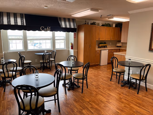 Small café/dining room with round tables and chairs, a kitchenette in the corner, wood floors and a striped awning labeled 'Cafe' over the windows.