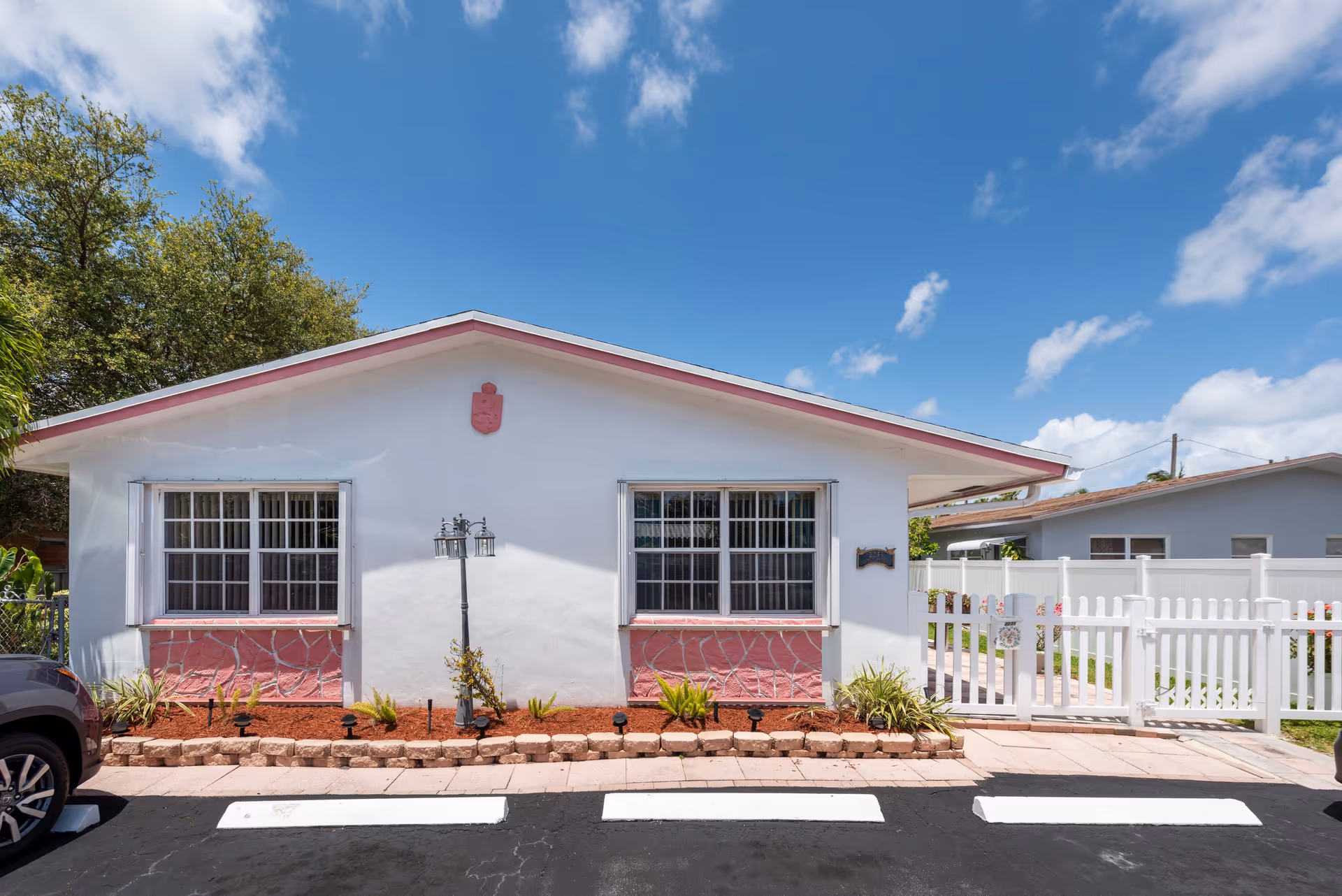 Exterior view of a single-story white building with two large windows, a small garden bed with plants and a lamp post in front, a white picket fence on the right side, and a parked car partially visible on the left under a blue sky with scattered clouds.