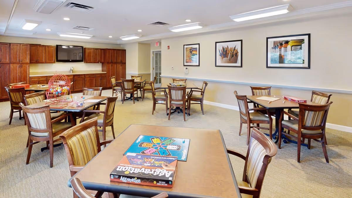 A well-lit activity room with multiple wooden tables and chairs arranged for group games. On the nearest table, there is a board game called Aggravation and a bingo cage with colorful balls. The room has beige walls adorned with framed pictures and a large flat-screen TV mounted above wooden cabinets.