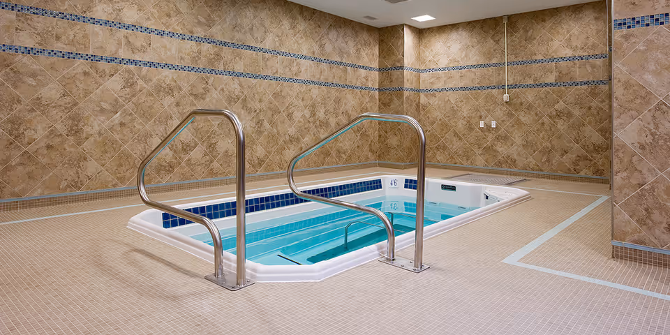 Indoor tiled therapy/spa pool with stainless handrails in a beige-tiled room.