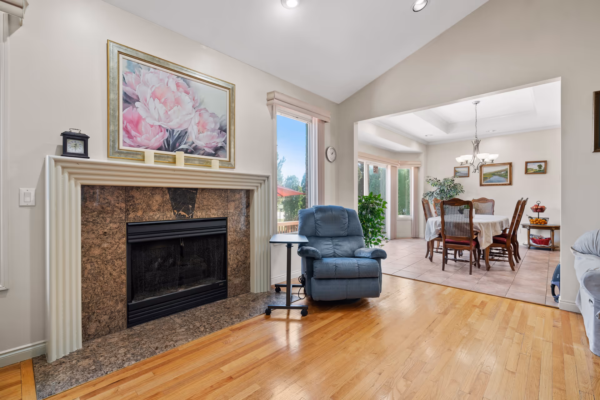 Interior view of a senior living facility showing a cozy living room with a blue recliner chair next to a fireplace with a floral painting above it. The room has hardwood floors and a large window letting in natural light. Adjacent to the living room is a dining area with a wooden dining table and chairs, a chandelier, and framed artwork on the walls.