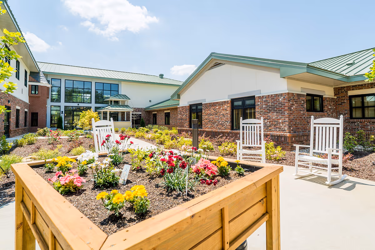 Sunny courtyard with raised wooden planters filled with flowers and white rocking chairs outside a brick senior living building.