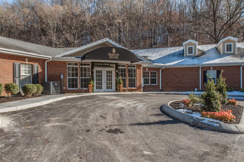 Front entrance of Barton House Memory Care, a single-story brick building with a covered entry, circular driveway, and landscaped islands.