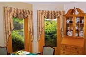 Interior view of a room with two tall windows framed by patterned curtains. Outside the windows, green foliage is visible. To the right, there is a wooden hutch displaying decorative plates and a wooden desk with drawers. A portion of a dining table and chairs is visible in the foreground.