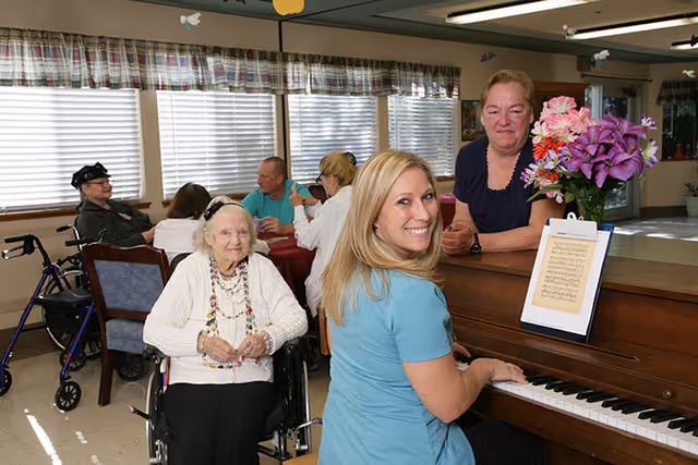 A group of elderly people and caregivers gathered in a common room. One woman in a blue shirt is playing a piano with sheet music and a vase of flowers on top. An elderly woman in a wheelchair and another woman stand nearby, smiling. Other people are seated at tables in the background.
