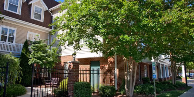 Exterior view of a senior living facility named The Willows, showing a brick and siding building partially shaded by green trees and bushes. There is a black metal fence with a gate, a bench, and a sidewalk leading to the entrance. The scene is bright and sunny.