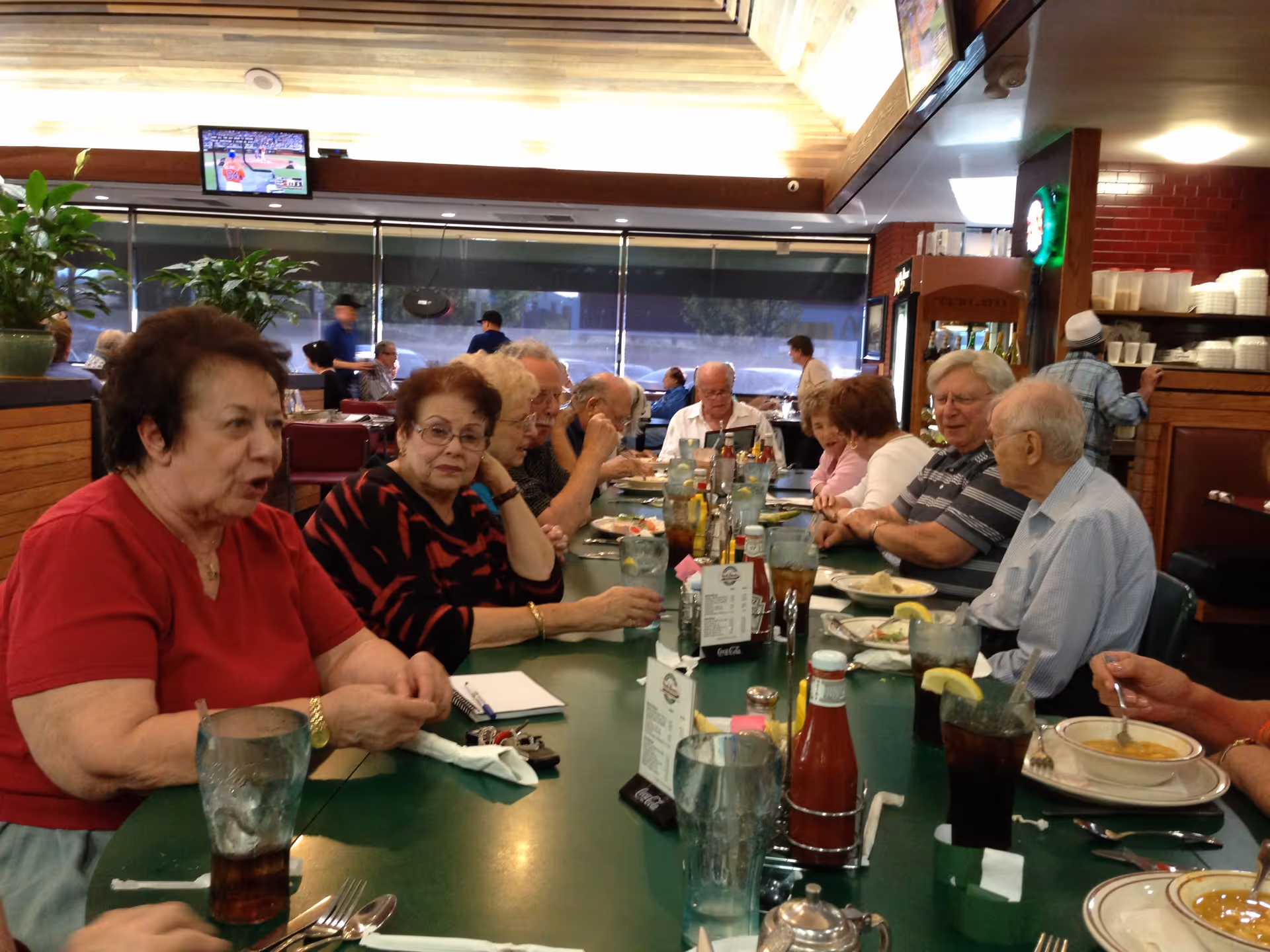 A group of elderly people sitting around a long dining table in a restaurant, engaged in conversation and eating. The table is set with glasses of iced drinks, plates of food, condiments, and menus. The background shows other diners and large windows.