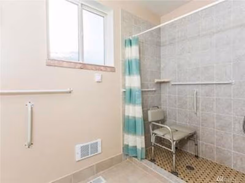 A bathroom with a walk-in tiled shower area featuring a metal shower chair and grab bars. The shower has a green and white striped curtain. There is a window above the shower area and additional grab bars on the adjacent wall.