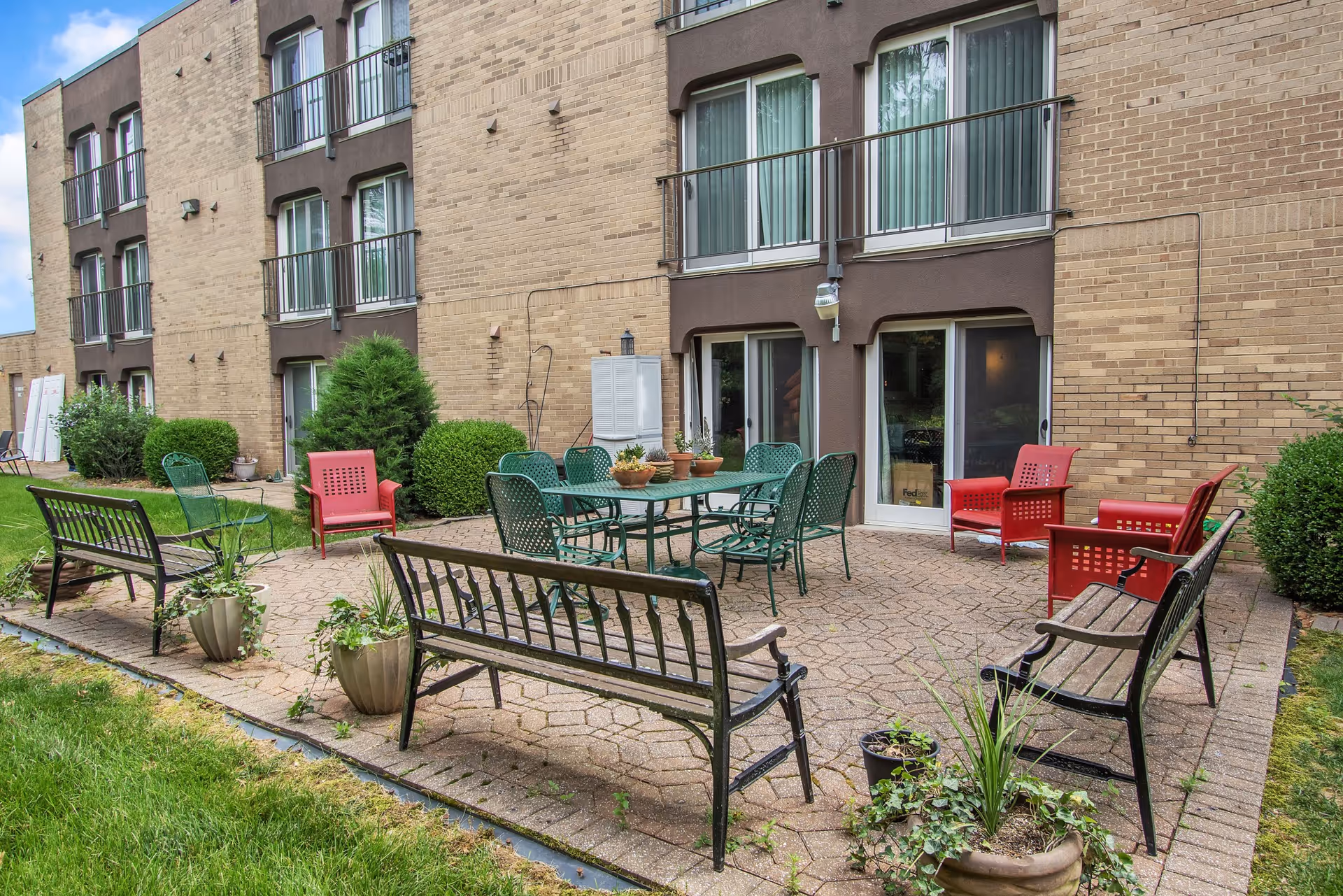 Outdoor patio courtyard with metal benches, a table and chairs, potted plants, and sliding doors along a brick apartment building.