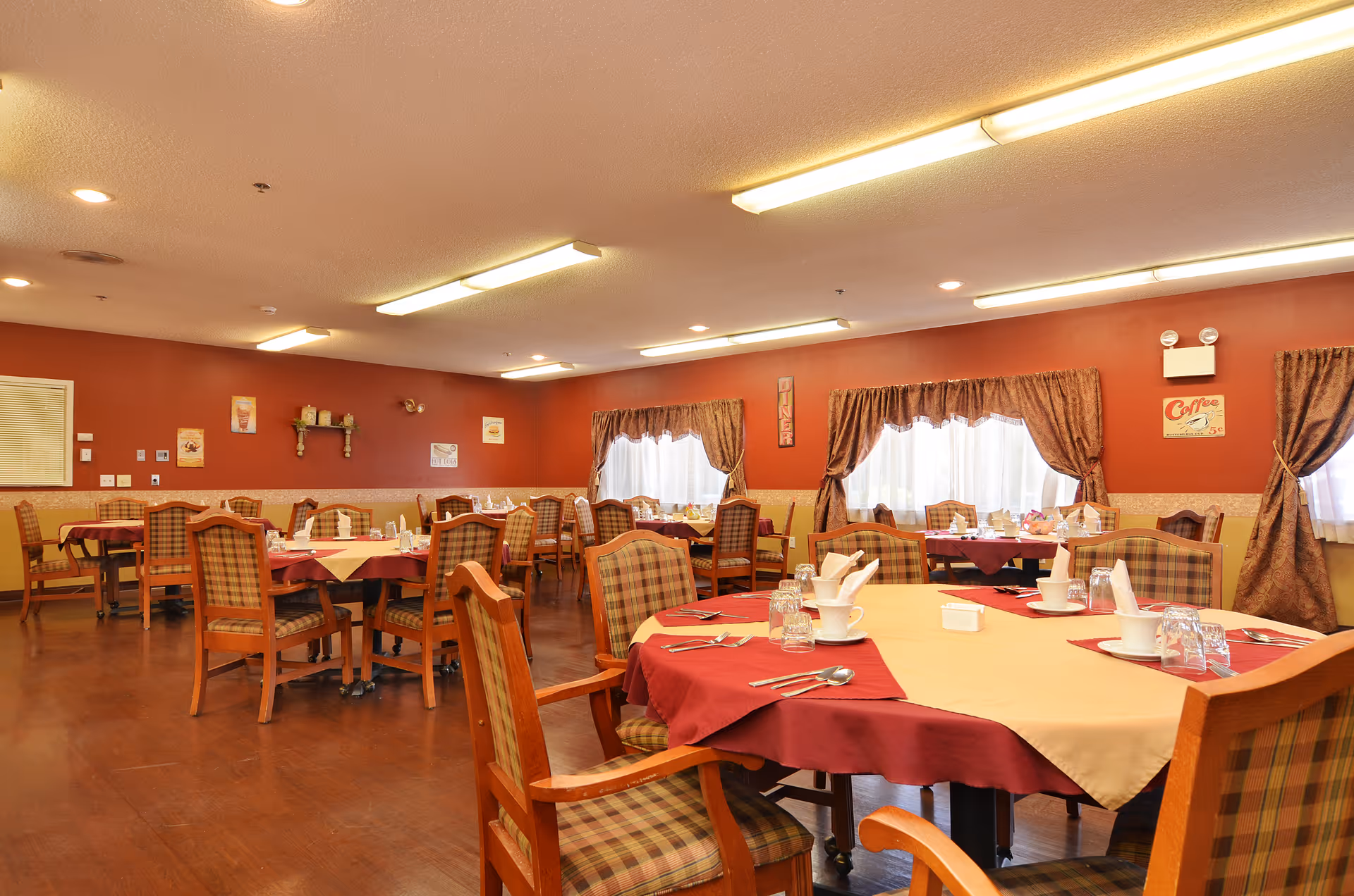 A dining room with multiple tables covered in red and beige tablecloths, set with cups, glasses, napkins, and cutlery. The room has wooden chairs with plaid upholstery, red walls, and windows with brown curtains letting in natural light. Various wall decorations and warm ceiling lights are visible.
