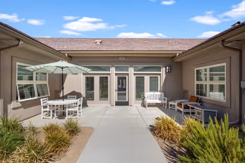 Outdoor patio area at The Monarch at Henderson with a round table and umbrella on the left, white bench seating on the right, surrounded by plants and shrubs under a blue sky with some clouds.