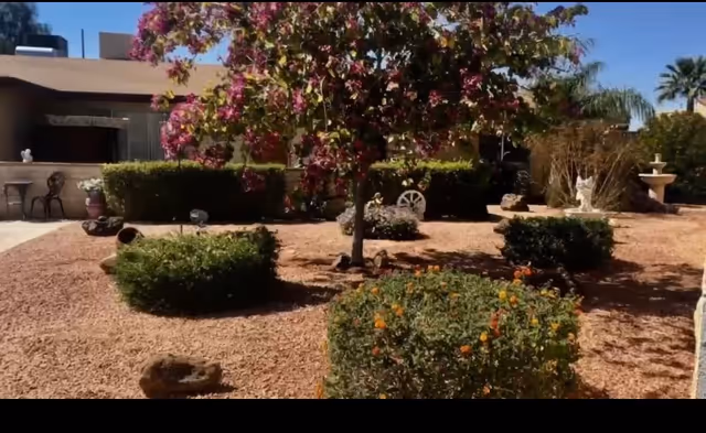 A landscaped outdoor garden area with flowering bushes, a tree with pink blossoms, and decorative elements including a white wagon wheel and a small statue. In the background, there is a single-story building with a patio area and outdoor seating.