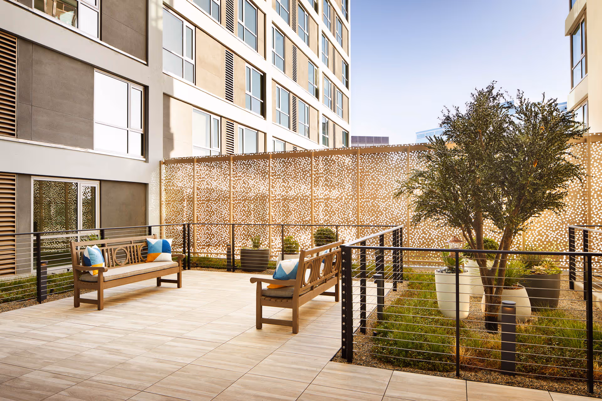 Outdoor patio area at a senior living facility with two wooden benches featuring colorful cushions, a decorative privacy screen, potted plants, and a small tree, surrounded by the building's exterior walls and windows.