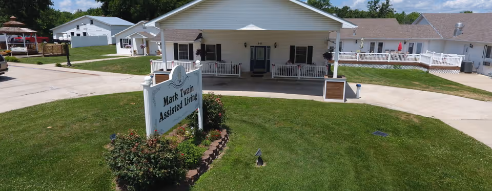 Front exterior view of Mark Twain Assisted Living facility showing a white building with a covered porch, surrounded by a well-maintained lawn and a sign with the facility's name in front.