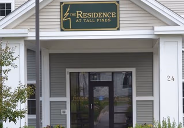 Entrance of a senior living facility named The Residence at Tall Pines, showing a covered porch with white pillars, a green sign with gold lettering, and the number 24 on the right pillar.