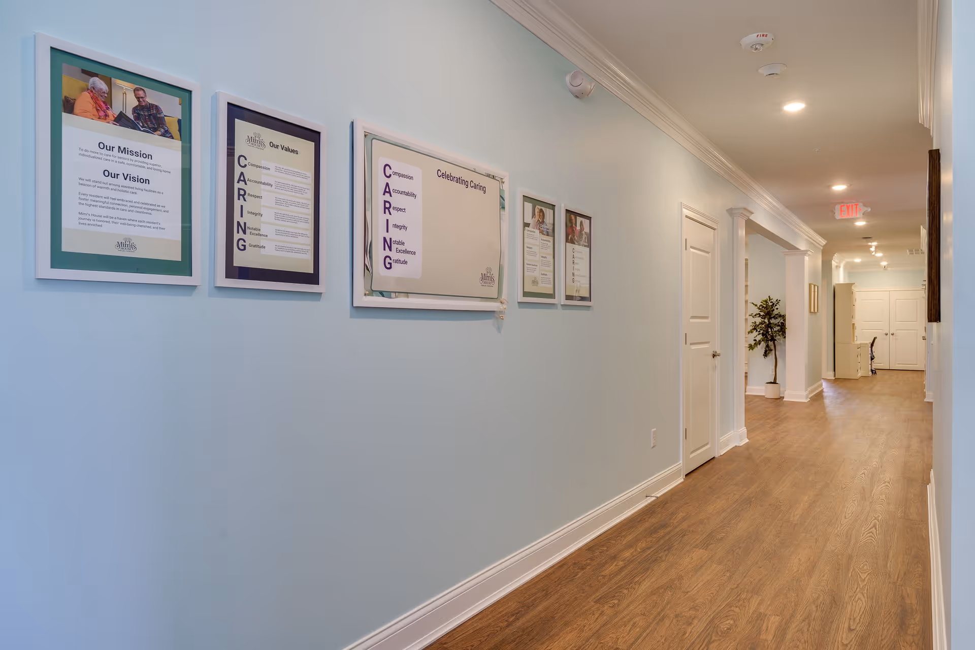 A clean, well-lit hallway in Mimi's House Assisted Living and Memory Care with light blue walls and wood flooring. Several framed posters and informational signs are displayed on the left wall, including the facility's mission, vision, and values. The hallway has white doors on the right side and a potted plant further down the corridor. Ceiling lights and an exit sign are visible at the far end.