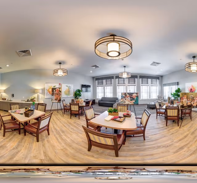 Bright communal activity/dining room with multiple tables and chairs, lounge seating by large windows, and pendant lights.
