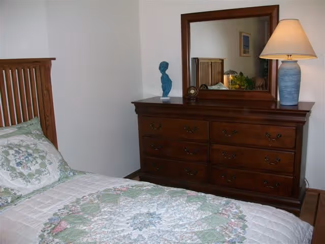 A bedroom with a wooden bed frame and a bed covered with a quilted bedspread featuring a floral and geometric pattern in green, white, and pink. Next to the bed is a wooden dresser with six drawers, a blue decorative bust, a small clock, and a blue lamp with a beige lampshade. A large mirror is mounted on the wall above the dresser, reflecting part of the bed and a framed picture on the opposite wall.
