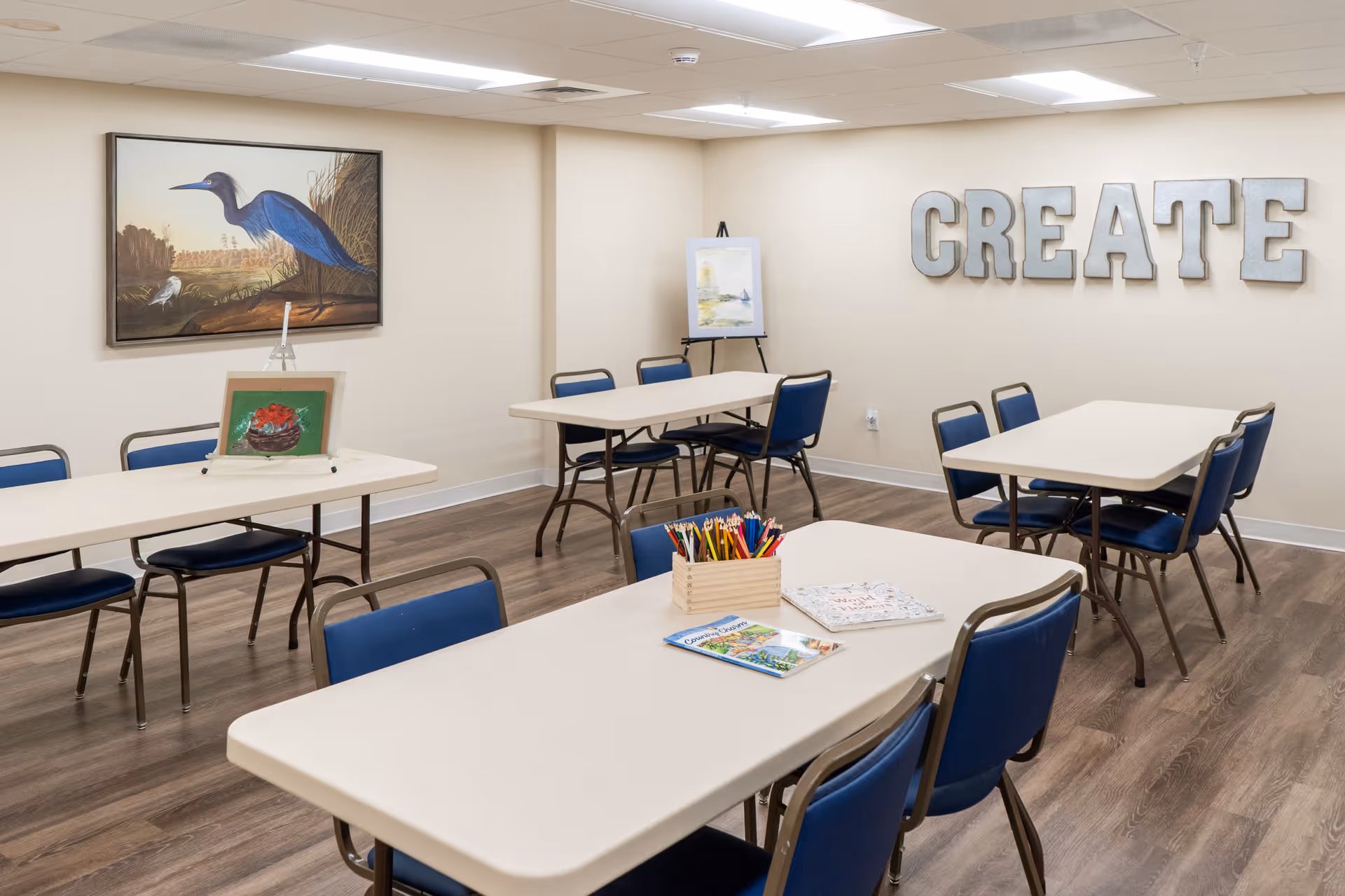A bright, clean activity room with several rectangular tables and blue chairs arranged around them. On the tables are art supplies including colored pencils and coloring books. The walls are decorated with a large painting of a blue heron and a smaller painting on an easel. Large metallic letters spelling 'CREATE' are mounted on one wall.