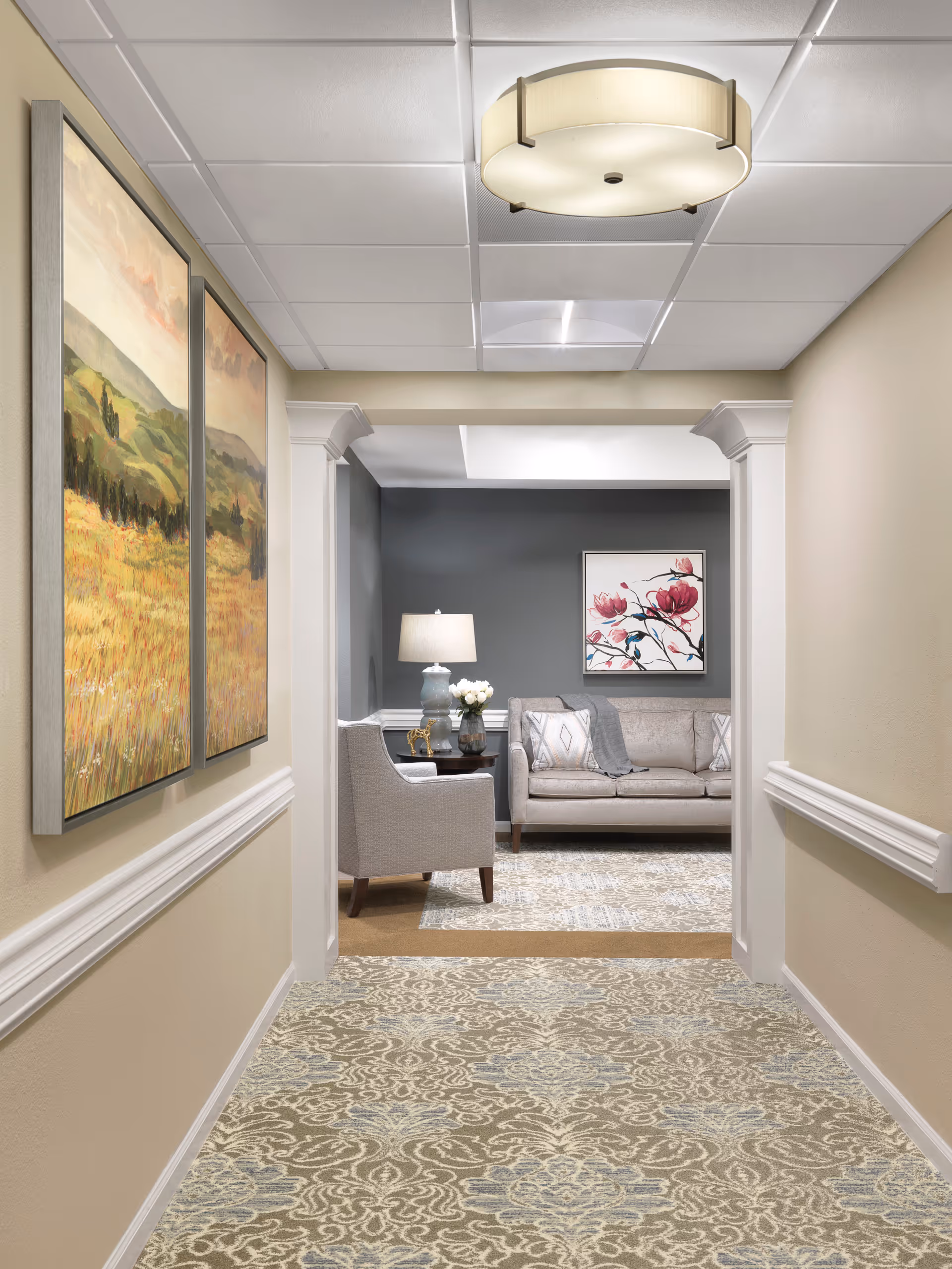 A hallway with patterned carpet and beige walls leads to a cozy sitting area with a gray sofa, an armchair, a side table with a lamp and flowers, and a framed floral artwork on a dark gray wall. Two landscape paintings hang on the left hallway wall, and a round ceiling light fixture is visible.