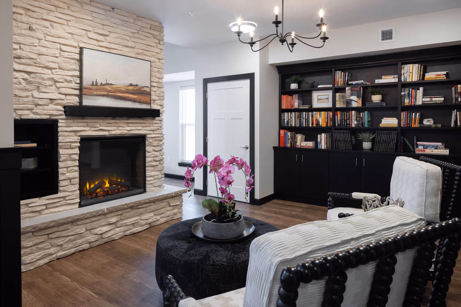 A cozy living room with a stone fireplace featuring a lit fire, a painting above the mantel, two white cushioned chairs with dark wooden frames, a round black ottoman with a potted orchid on top, and a large black bookshelf filled with books and decorative plants. The room has wooden flooring and a modern chandelier hanging from the ceiling.