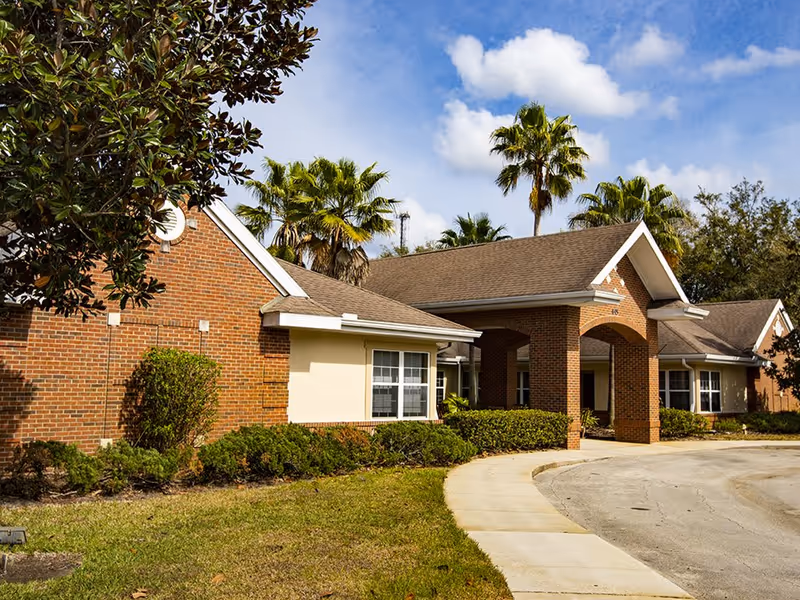 Exterior view of a single-story brick building with a covered entrance, surrounded by palm trees and shrubs under a partly cloudy blue sky.