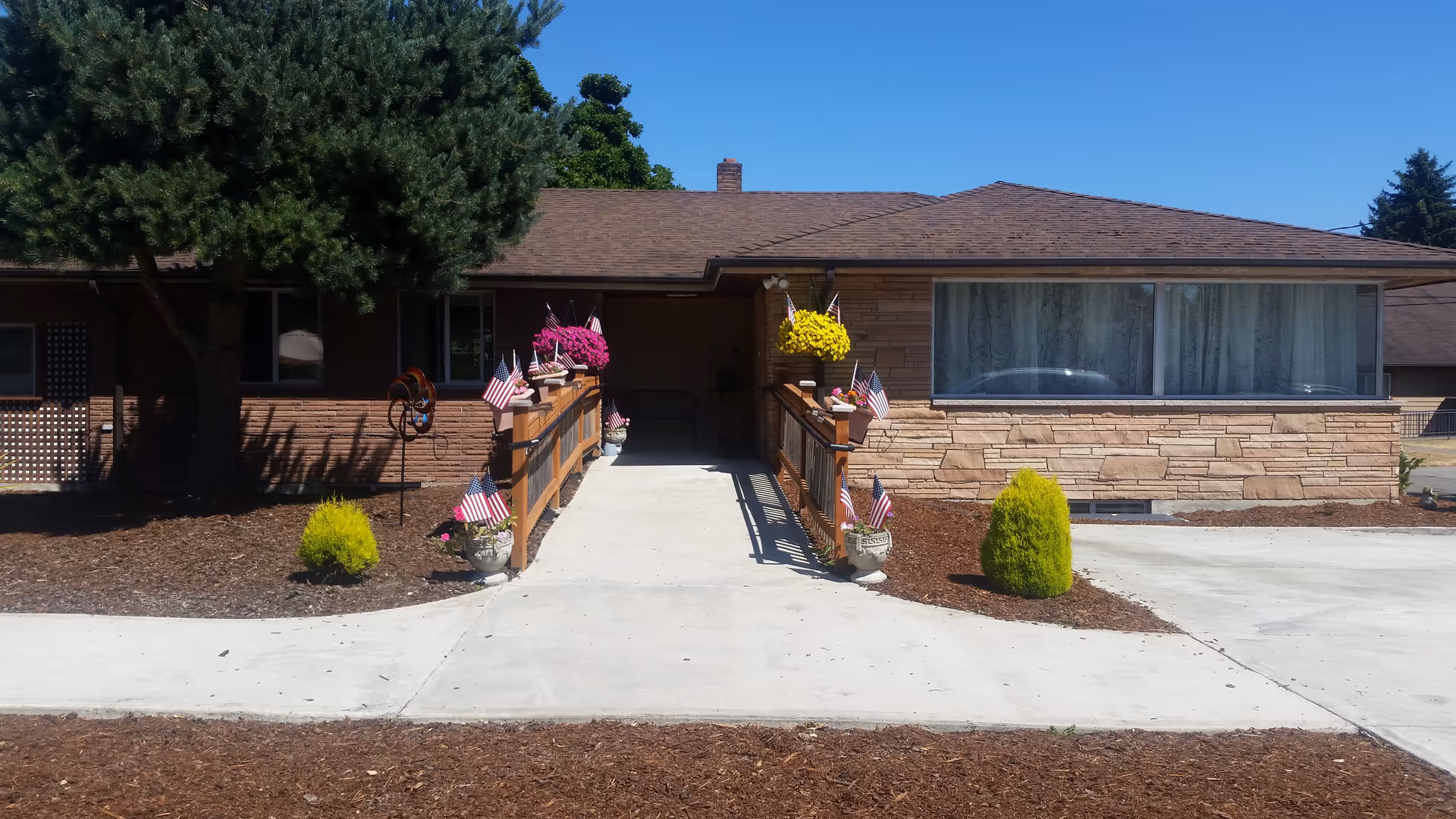 Front exterior view of a single-story building with a ramp leading to the entrance. The ramp is decorated with small American flags and flower pots with pink and yellow flowers. There are small shrubs and a large tree on the left side of the building. The building has a stone facade and a large window with curtains.