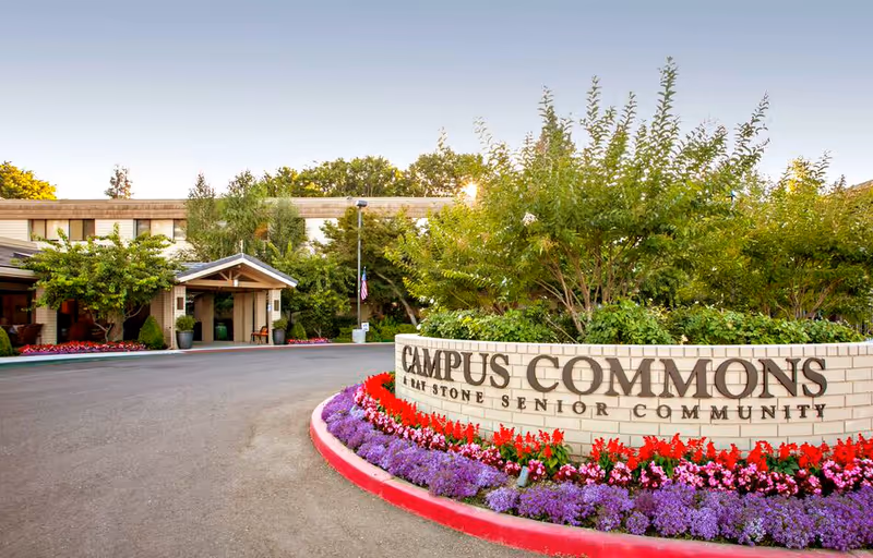 Entrance of Campus Commons, a senior living community, featuring a curved brick sign with the community name surrounded by colorful flowers and greenery, with the building and trees in the background under a clear sky.