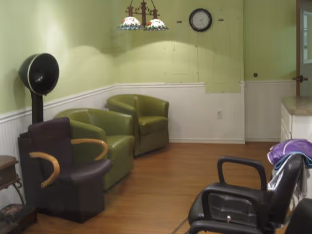 Interior room with light green walls and wood flooring featuring two green armchairs, a black salon hair dryer chair, a black chair with armrests, a small side table, a wall clock, and a hanging light fixture with stained glass shades.