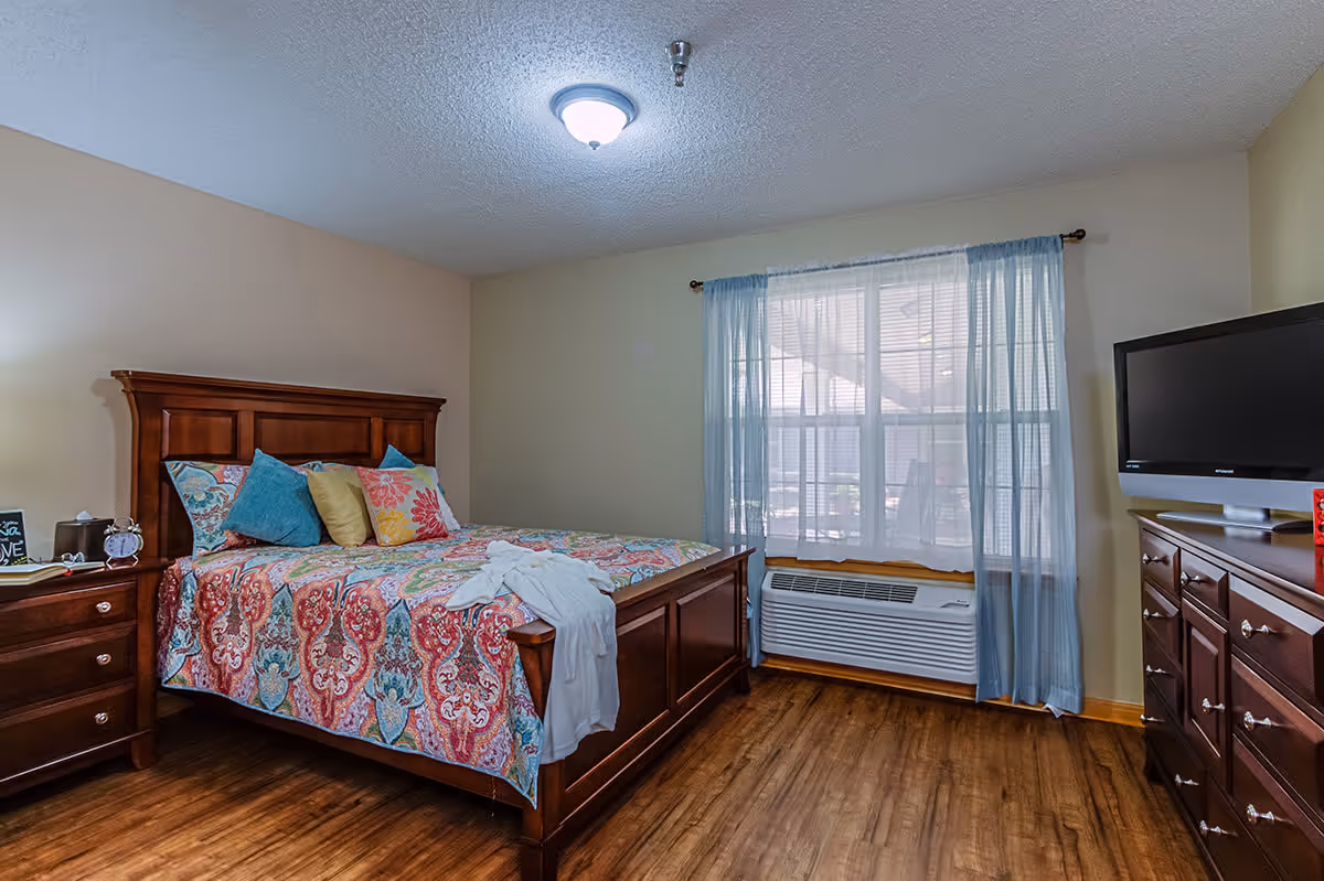 A cozy bedroom with a wooden bed frame and colorful patterned bedding, several pillows, a nightstand with a clock and tissue box, a window with sheer blue curtains, a wall-mounted air conditioning unit below the window, and a wooden dresser with a flat-screen TV on top.