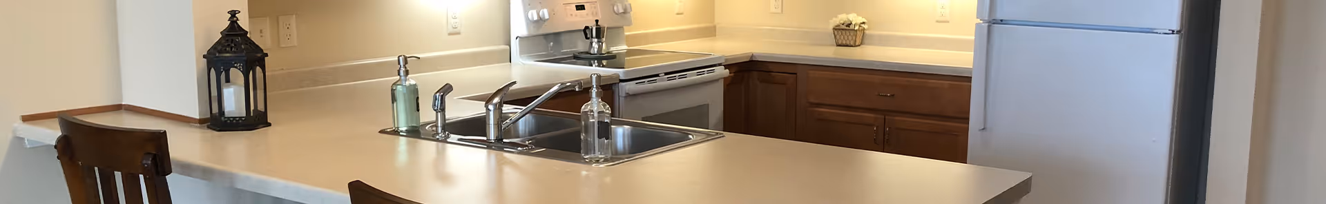 A modern kitchen area with a beige countertop island featuring a double sink and a soap dispenser. Behind the island, there is a white electric stove with a pot on it, wooden cabinets, and a small decorative basket on the counter. A black lantern is placed on the left side of the island, and two wooden chairs are partially visible.